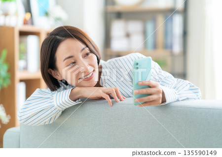 A middle-aged woman lying on the sofa in the living room and looking at her smartphone 135590149