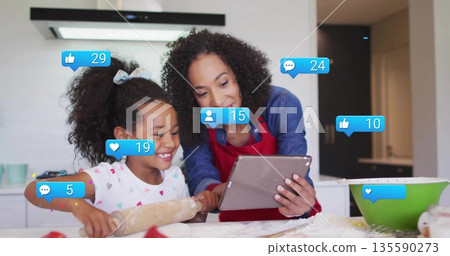Mother wearing red apron guiding daughter pressing dough with tablet in kitchen, with social icons Mother wearing red apron guiding daughter pressing dough with tablet in kitchen, with social icons 135590273