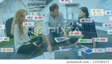 Collaborating team arranging charts photos around laptop on office floor, overlaying social metrics 135590337