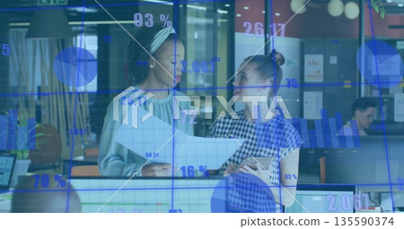 Professional women reviewing printed data charts and tablet in office with monitors, data visuals Professional women reviewing printed data charts and tablet in office with monitors, data visuals 135590374