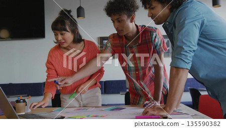 Pointing and discussing printed layouts around modern meeting table with laptop, sticky notes Pointing and discussing printed layouts around modern meeting table with laptop, sticky notes 135590382