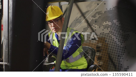Operating Korean forklift operator steering controls in warehouse, wearing hard hat and safety vest 135590411