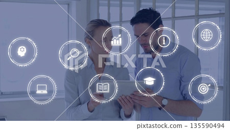 Standing business professionals viewing tablet screen in modern office, with digital overlay icons Standing business professionals viewing tablet screen in modern office, with digital overlay icons 135590494