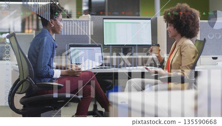 Reviewing data while two women holding coffee cups at office desk, dual monitors displaying graphs 135590668