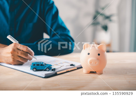 Man signs car finance contract near pink piggy bank as money savings for auto insurance and lease documents, using price management to save for buying vehicle on wooden office desk Man signs car finance contract near pink piggy bank as money savings for auto insurance and lease documents, using price management to save for buying vehicle on wooden office desk 135590929