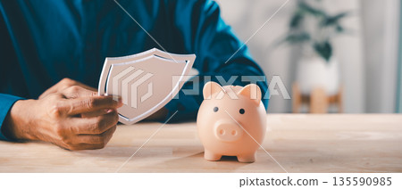 Man holds white shield icon near pink piggy bank to protect money, saving plan financial finance insurance saving shield concept ensures secure investment growth plus future safety Man holds white shield icon near pink piggy bank to protect money, saving plan financial finance insurance saving shield concept ensures secure investment growth plus future safety 135590985