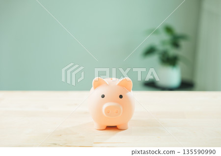 Pink piggy bank for money saving goals sits on wooden table, financial investment concept for personal finance, background showing blurred houseplant and minimalist home interior 135590990