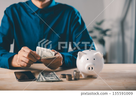 Man counts cash for digital savings mobile payment bank, piggy bank and phone help save online using smartphone pig app, holding dollar bills near stacks of coins on wooden table Man counts cash for digital savings mobile payment bank, piggy bank and phone help save online using smartphone pig app, holding dollar bills near stacks of coins on wooden table 135591009