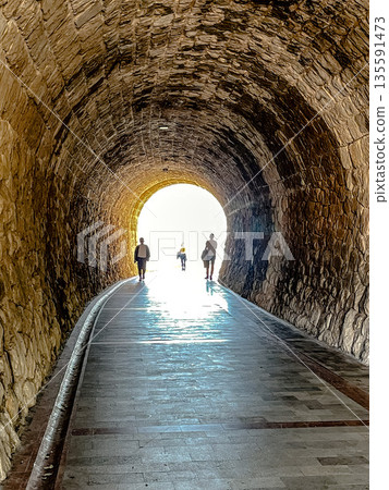 three individuals traverse shadowed pathway, people moving towards glowing tunnel entrance three individuals traverse shadowed pathway, people moving towards glowing tunnel entrance 135591473