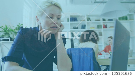 Leaning blonde-haired woman in navy blouse focusing on desktop monitor in open office, copy space 135591686