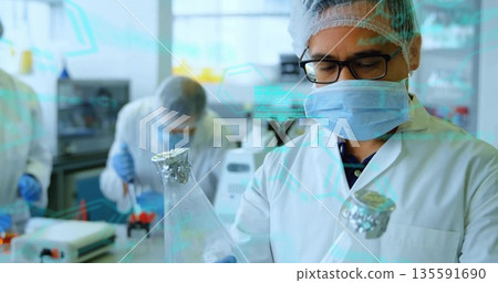 Mid adult scientist wearing lab coat holding Erlenmeyer flasks in lab, with pipette and centrifuge 135591690