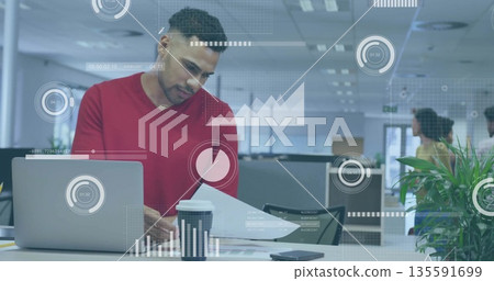 Man in red long-sleeve shirt reviewing papers in open-plan office, with laptop and data overlays Man in red long-sleeve shirt reviewing papers in open-plan office, with laptop and data overlays 135591699