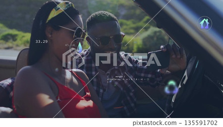 Smiling couple wearing sunglasses capturing selfie in beige convertible on rural road, smartphone 135591702