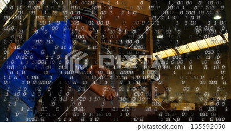 Operating machinist wearing ear protection adjusting lathe controls in workshop, binary overlay 135592050