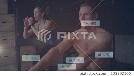 Rowing shirtless man and woman in blue tank top jumping in fitness studio, ergometer, notifications 135592100
