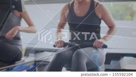 Rowing fitness partners pulling handles on rowing machines in gym cardio area with digital displays 135592104