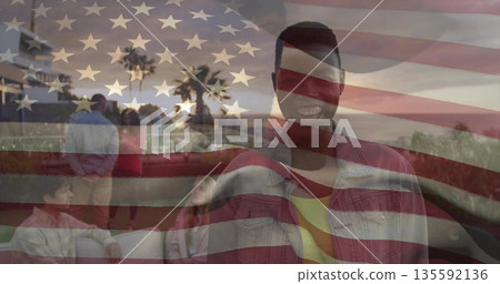 Smiling man wearing denim jacket standing on promenade at sunset with railing, palms, flag overlay 135592136