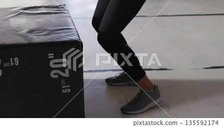 Squatting woman in black leggings on gym floor lines interacting with 20" 51 cm plyometric box Squatting woman in black leggings on gym floor lines interacting with 20" 51 cm plyometric box 135592174