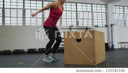 Woman bending knees preparing box jump in gym in sportswear, with box, kettlebells, ropes, table Woman bending knees preparing box jump in gym in sportswear, with box, kettlebells, ropes, table 135592190