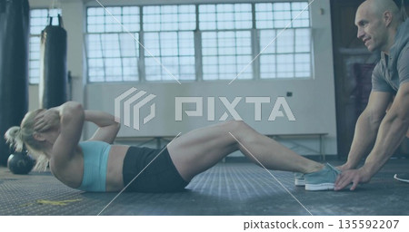 Performing sit-ups in sportswear, client with instructor holding feet in gym with punching bag Performing sit-ups in sportswear, client with instructor holding feet in gym with punching bag 135592207