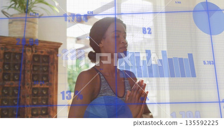Meditating woman holding palms at chest in studio with cabinet, potted plant and data overlays Meditating woman holding palms at chest in studio with cabinet, potted plant and data overlays 135592225