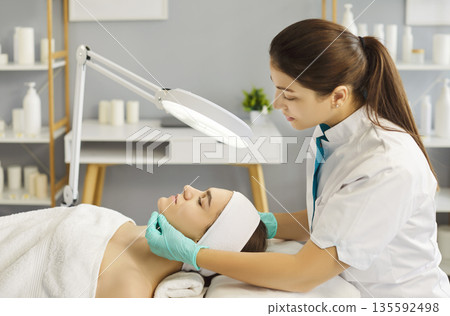 Cosmetologist examining client's face under magnifying lamp 135592498