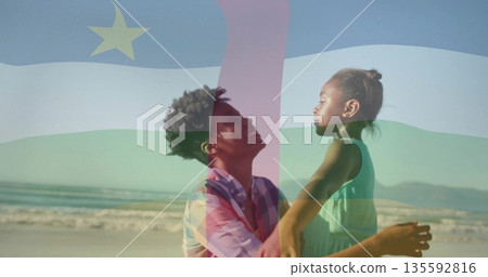 Smiling mother holding daughter at waist level on sandy beach, with semi-transparent flag overlay 135592816
