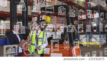 Reviewing warehouse staff checking stock on clipboard beside orange forklift, digital overlay icons 135592875