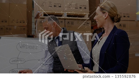 Colleagues pointing and reviewing logistics on whiteboard in warehouse, with laptop and box stacks Colleagues pointing and reviewing logistics on whiteboard in warehouse, with laptop and box stacks 135592876