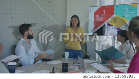 Presenting woman wearing suit gesturing at screen in conference room, with laptops, coffee cups 135592933