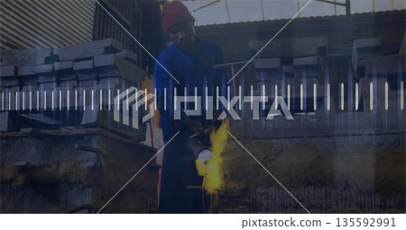 Worker wearing gloves pouring molten metal from crucible with tongs at foundry, with stacked molds 135592991
