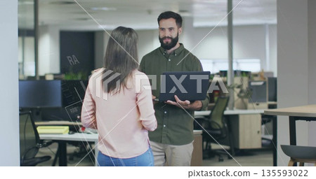 Discussing work two coworkers holding laptop while conversing in office, with desks, plant, stool Discussing work two coworkers holding laptop while conversing in office, with desks, plant, stool 135593022