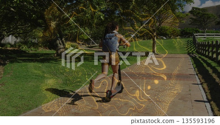 Woman wearing shorts riding electric scooter down park brick path, denim backpack, golden lights 135593196