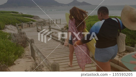 Walking friends in beachwear on beach boardwalk, carrying yellow surfboard, straw hat and tote bag Walking friends in beachwear on beach boardwalk, carrying yellow surfboard, straw hat and tote bag 135593205