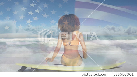 Sitting woman in orange swimsuit gazing at ocean on shore, with yellow surfboard and flag-overlay 135593301