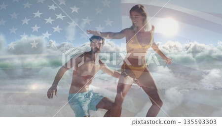 Riding surfboard on ocean wave, couple in swimwear with US flag and clouds overlay 135593303