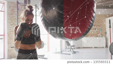 Punching female athlete wearing sportswear striking heavy bag in fitness studio, with boxing gloves Punching female athlete wearing sportswear striking heavy bag in fitness studio, with boxing gloves 135593311