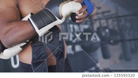Wrapping shirtless boxer securing bright blue hand wrap at boxing gym, with gloves and bags 135593320