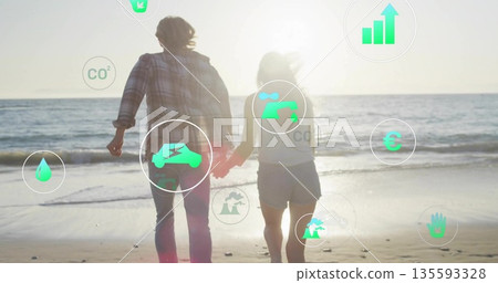 Walking couple holding hands toward ocean at sandy shore, with floating eco and financial icons 135593328