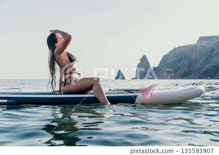 Woman Paddleboard Ocean Coastline - A woman in a bikini sits on a paddleboard in the ocean, looking out at a rocky coastline. The sun is shining and the water is calm. Woman Paddleboard Ocean Coastline - A woman in a bikini sits on a paddleboard in the ocean, looking out at a rocky coastline. The sun is shining and the water is calm. 135593907