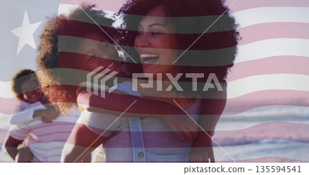 Carrying woman giving piggyback ride along sandy beach with waves at sunset, American flag overlay 135594541