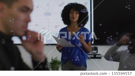 Presenting woman wearing blue blouse and pants holding papers in meeting room, with network display Presenting woman wearing blue blouse and pants holding papers in meeting room, with network display 135594557