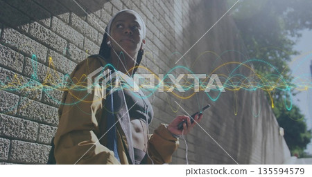 Leaning sporty woman holding smartphone listening through earphones on sunlit sidewalk, sound waves 135594579