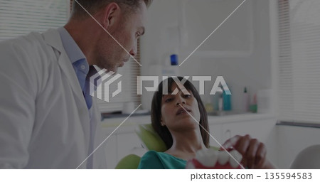 Discussing treatment dentist in lab coat, patient wearing teal gown holding dental model at clinic Discussing treatment dentist in lab coat, patient wearing teal gown holding dental model at clinic 135594583
