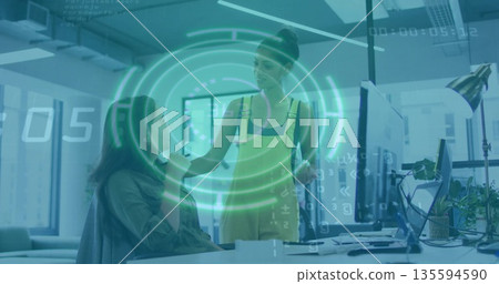 Collaborating woman and coworker visualizing data on monitors in office, with holographic overlay 135594590