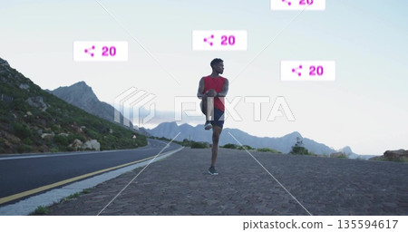 Bending adult male touching toes on turnout, with asphalt road, rocky hillside and mountain peaks 135594617