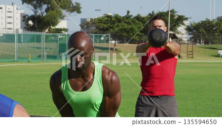 Men wearing neon tank top and red shirt swinging black kettlebells on grassy sports field 135594650