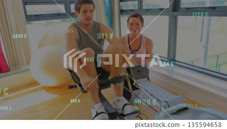 Rowing personal trainer and client on rower in gym wearing sportswear with exercise ball, tablet Rowing personal trainer and client on rower in gym wearing sportswear with exercise ball, tablet 135594658