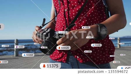 Adjusting female photographer holding DSLR camera on boardwalk, with floating social media icons Adjusting female photographer holding DSLR camera on boardwalk, with floating social media icons 135594672