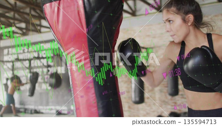 Punching female boxer wearing sports bra and gloves training in boxing gym, with heavy punching bag 135594713